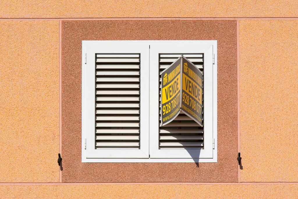 White shutter window with a for sale sign on peach building exterior.