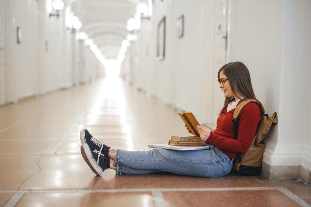 A young woman in casual attire sits reading in a well-lit hallway.