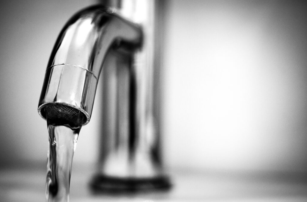 Black and white close-up of water flowing from a shiny stainless steel faucet indoors.