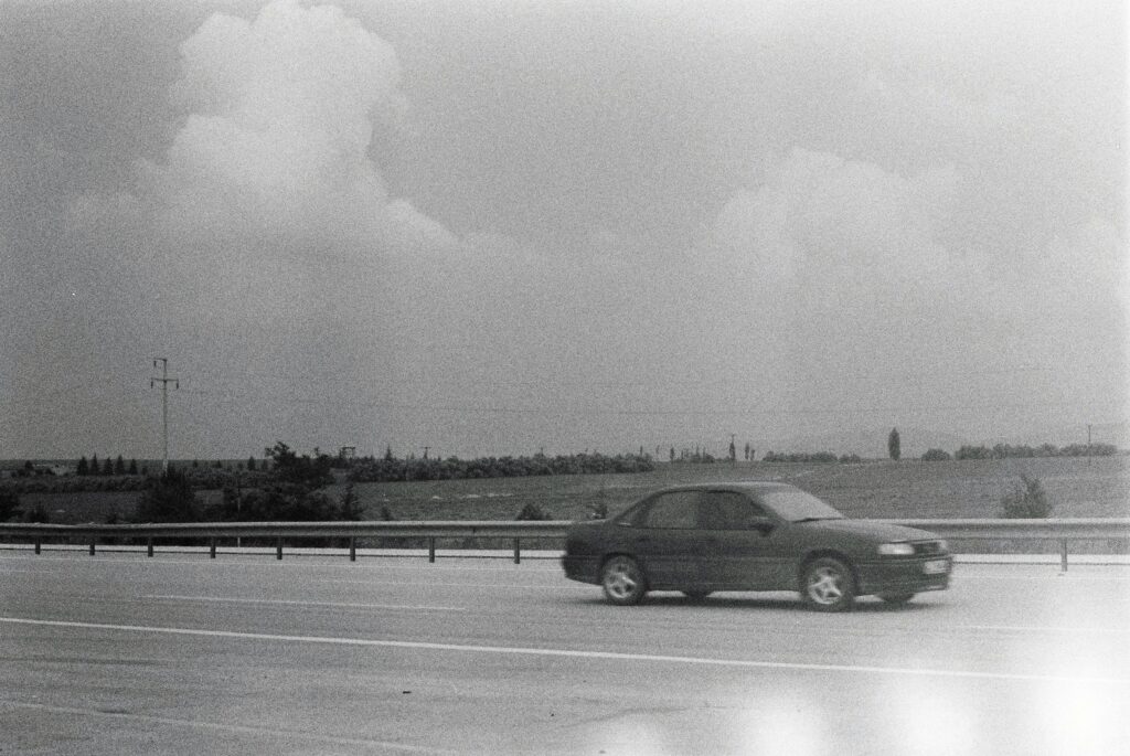 A vintage black and white photo of a car on a rural road under a cloudy sky.
