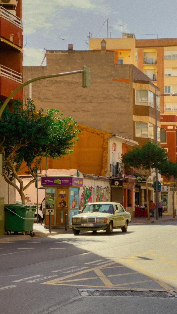A vintage car parked on a lively urban street lined with colorful buildings under a bright sky.
