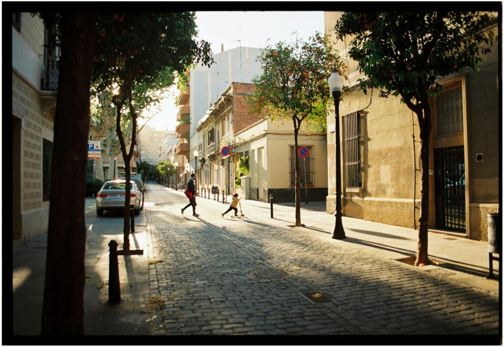 A child and an adult stroll down a cobblestone street in Barcelona, surrounded by trees and urban architecture.