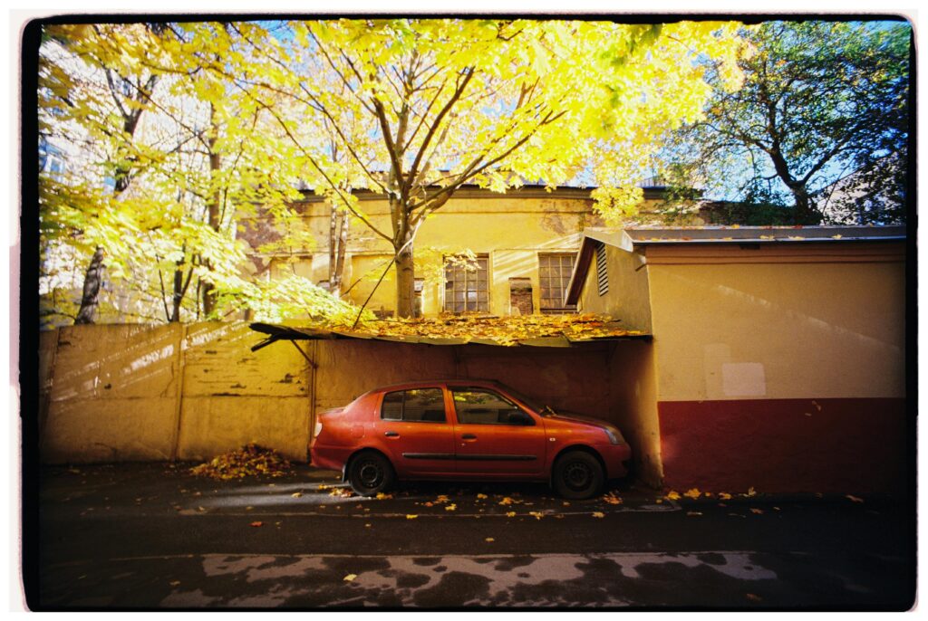 A red car parked under a tree with bright yellow leaves in autumn.